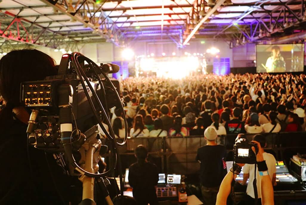 ライブ会場でステージ後方からカメラで撮影するスタッフ